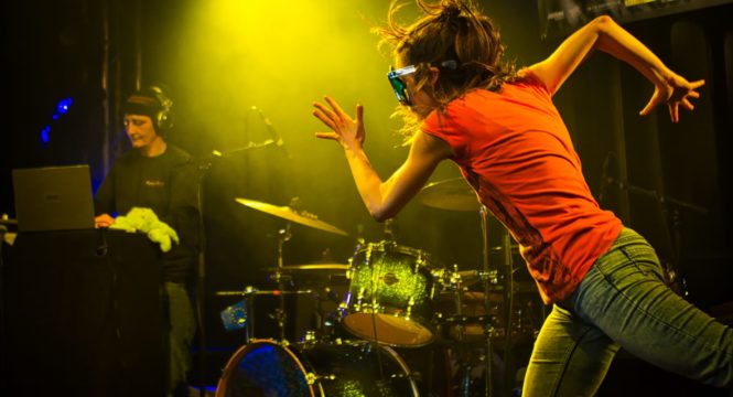 Eine Frau die tanzt, von der Seite fotografiert. Sie streckt einen Arm nach vorne und einen nach hinten aus, als würde sie rennen. Sie trägt ein rotes T-Shirt und eine blaue Jeans, dazu eine Sternsonnenbrille. Im Hintergrund sind ein Schlagzeug und eine DJane zu sehen.