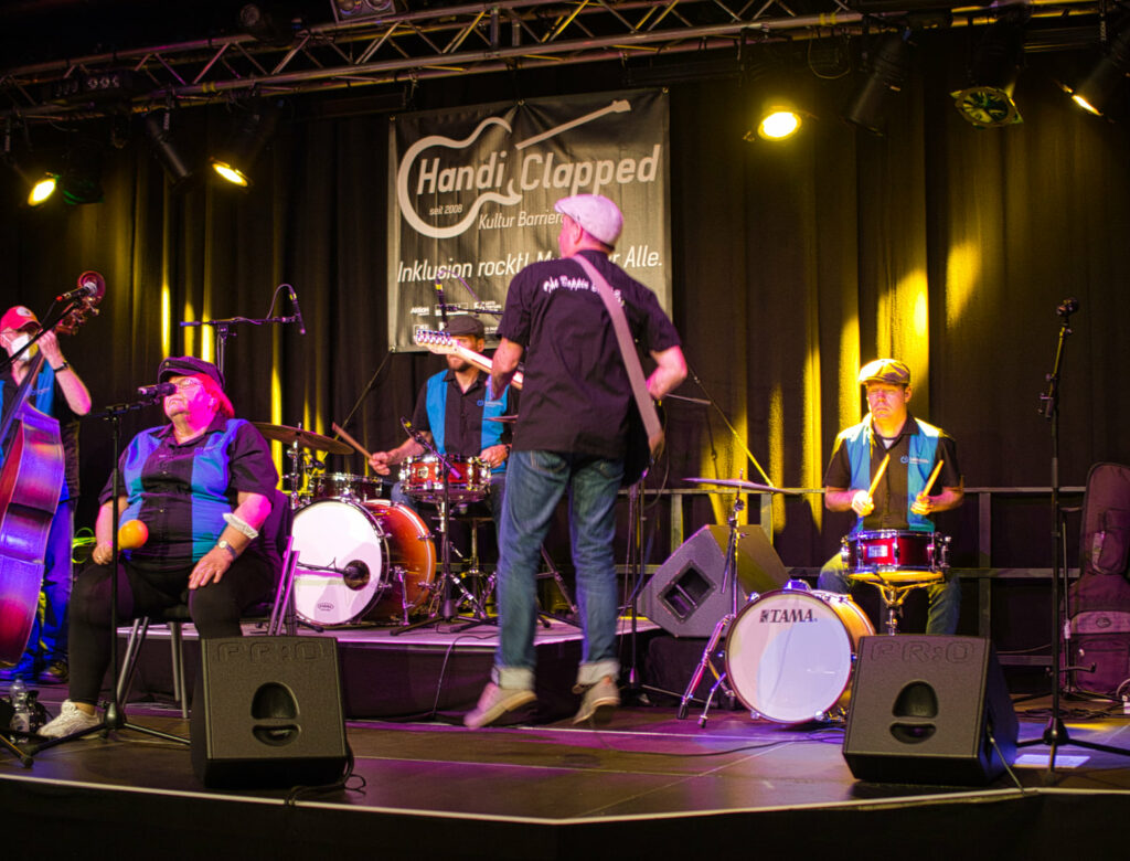 Eine Band in einheitlichen blau schwarzen Hemden auf der Bühne. Ein E-Gittarist ist mitten im Luftsprung in der Rückansicht, zwei Schlagzeuger und eine sitzende Sängerin im Hintergrund