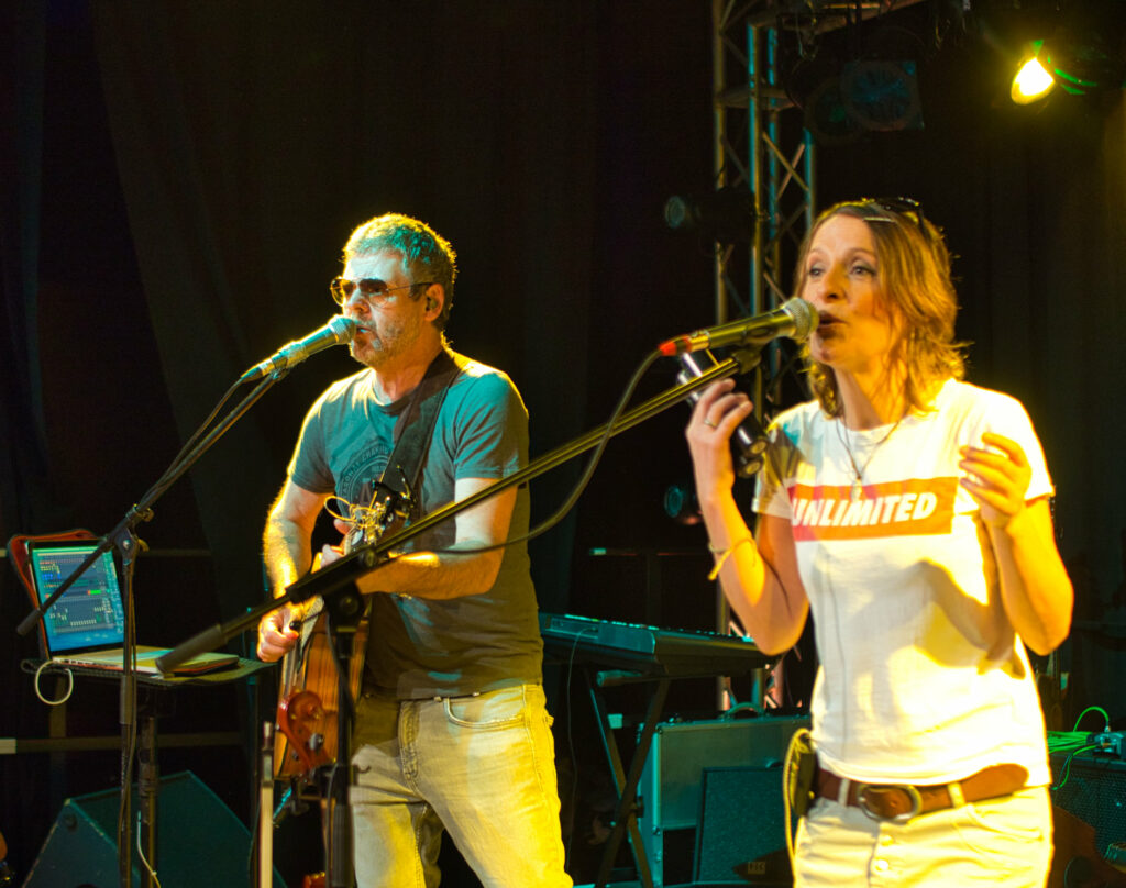 Ein Sänger mit E-Gitarre und eine Sängerin mit Klanghölzern. Beide Sagen ins Mikrofon, auf einer Bühne innen. Der Hintergrund dunkel.