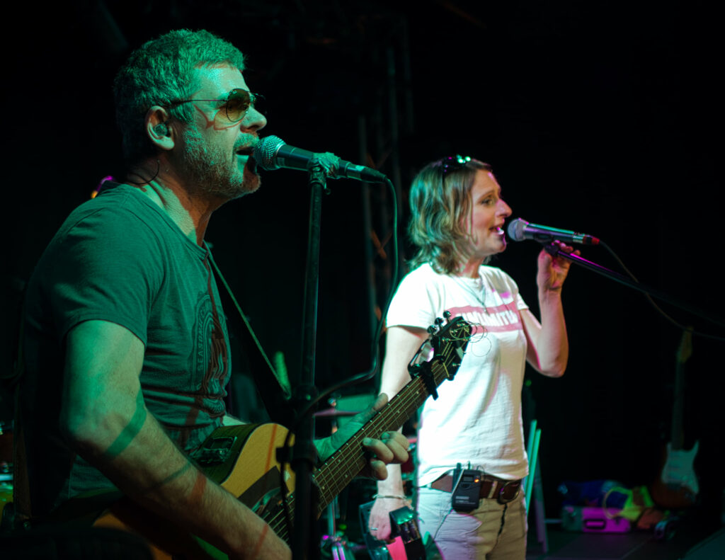Der gleiche Mann mit Gitarre und Sonnenbrille im Vordergrund, die Frau im Hintergrund, beide singen ins Mikrofon