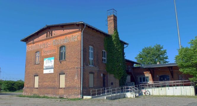 Ein bild von einer alten Brauerei aus Ziegelsteinen, alleinstehend, oben ist noch "Schultheiss" zu lesen, darüber blauer Himmel, vor dem Gebäude eine Rollstuhlrampe für barrierefreien Zugang.