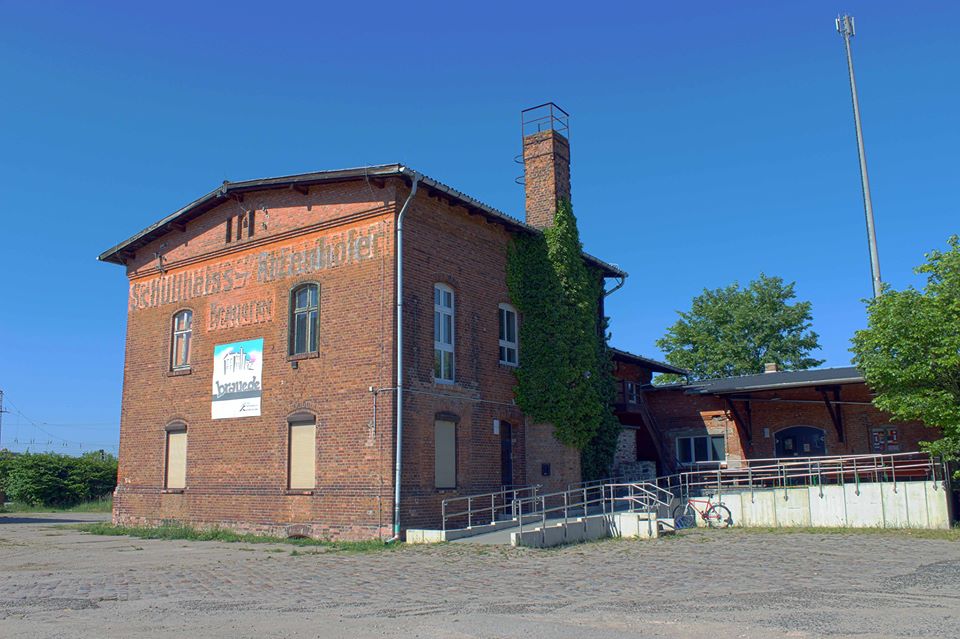 Ein bild von einer alten Brauerei aus Ziegelsteinen, alleinstehend, oben ist noch "Schultheiss" zu lesen, darüber blauer Himmel, vor dem Gebäude eine Rollstuhlrampe für barrierefreien Zugang.