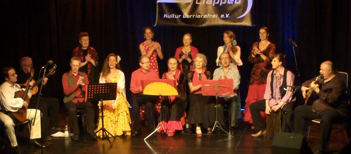 13 Chormitglieder und drei Gitarrenspieler mit Akustikgitarre live beim Auftritt auf der Bühne. Sie tragen Flamenco Trachten. Die Hälfte steht und die andere sitzt davor auf Stühlen in einer Reihe