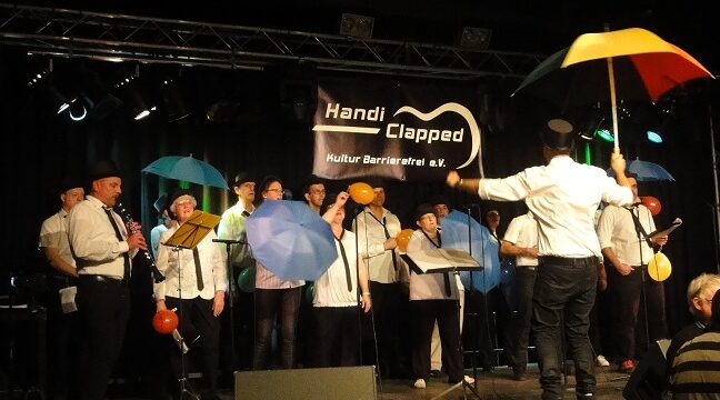 Ein Chor mit 16 Mitgliedern live auf der Bühne. Sie tragen alle weiße Hemden und schwarze lange hosten. 5 von ihnen halten bunte Regenschirme hoch
