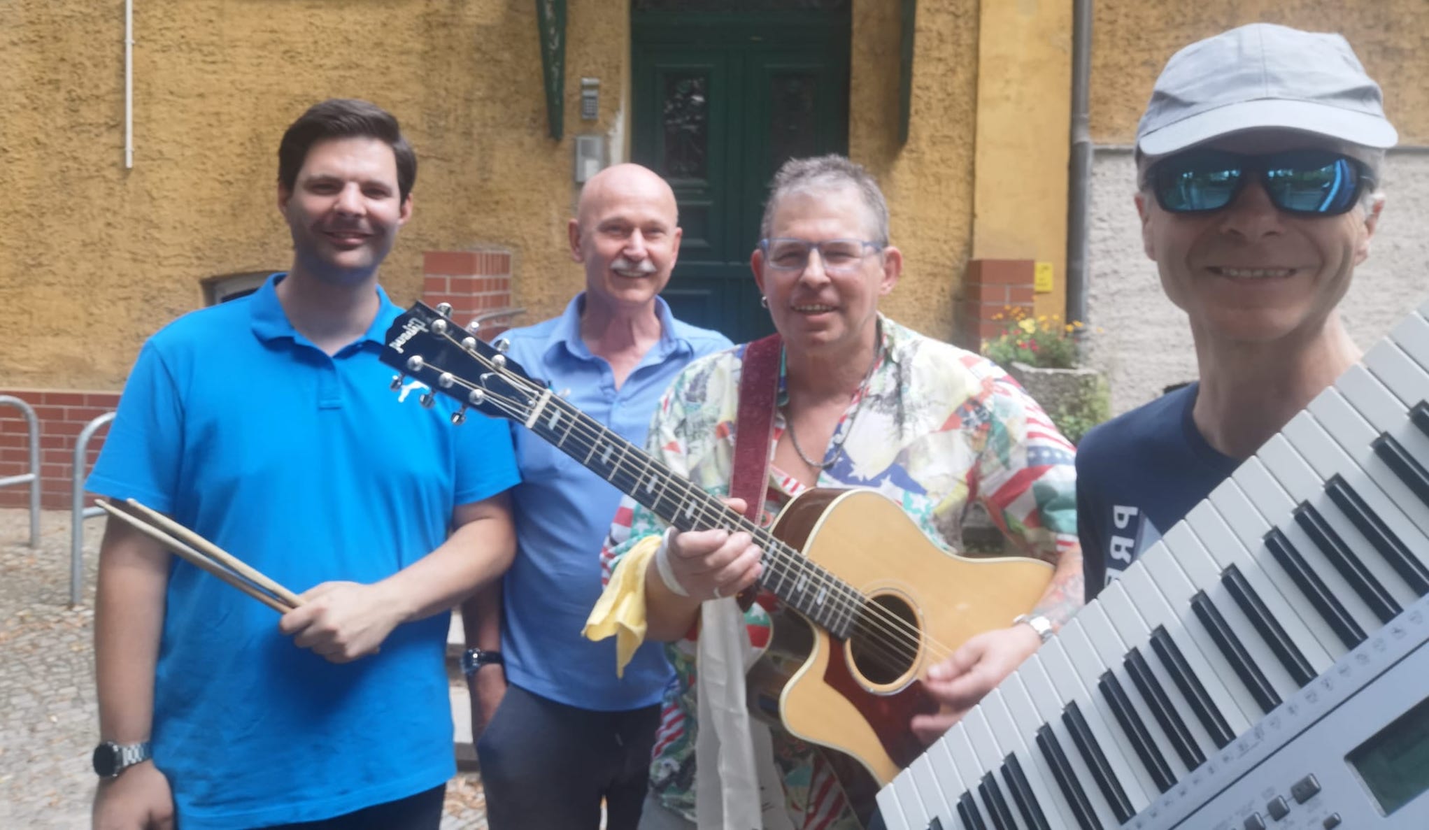 Vier Musiker vor einer Haustür, mit Schlagzeugstöcken, Gitarre und Keyboard