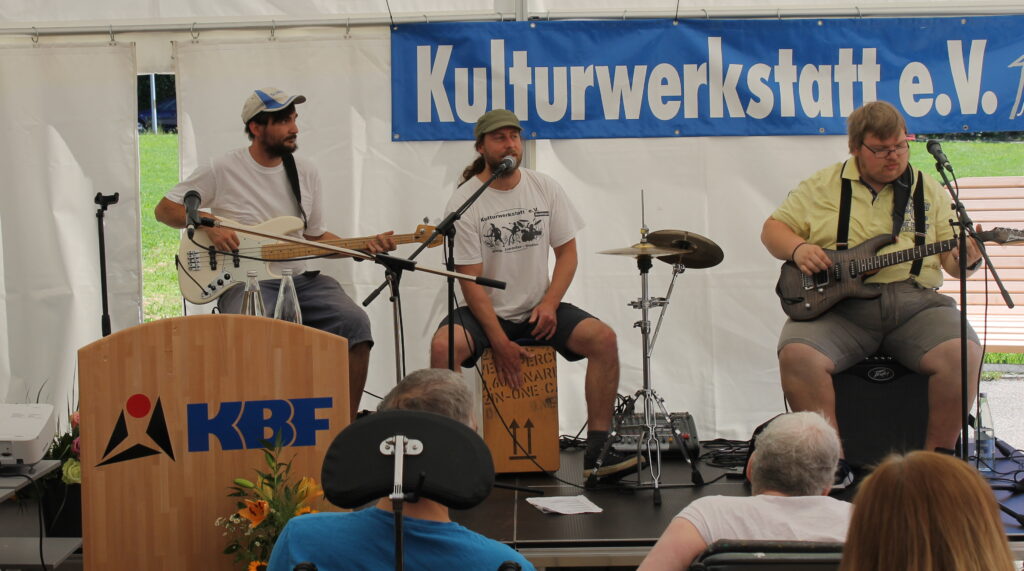 Drei Männer auf der Bühne machen Musik mit Gitarre, Zachen und Bass, im Hintergrund ein Banner "Kulturwerkstatt e.V."