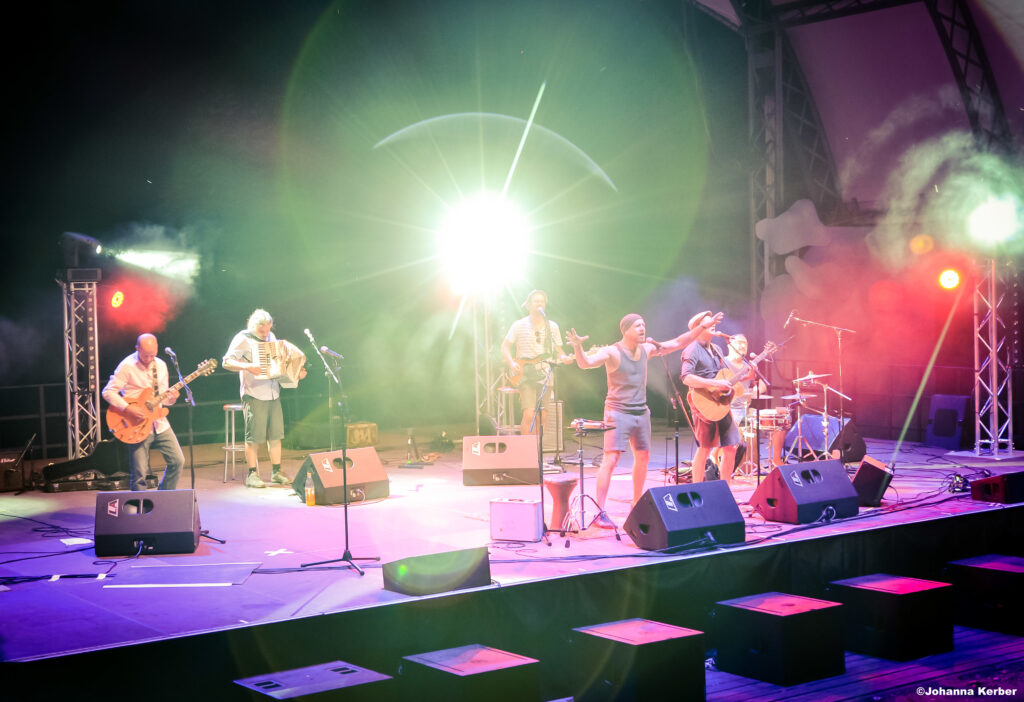 Buntfoto von 6 Musikern die Live mit E-Gitarre, Akkordeon, Gesang, Schlagzeug, Bass und Akustikgitarre auftreten.