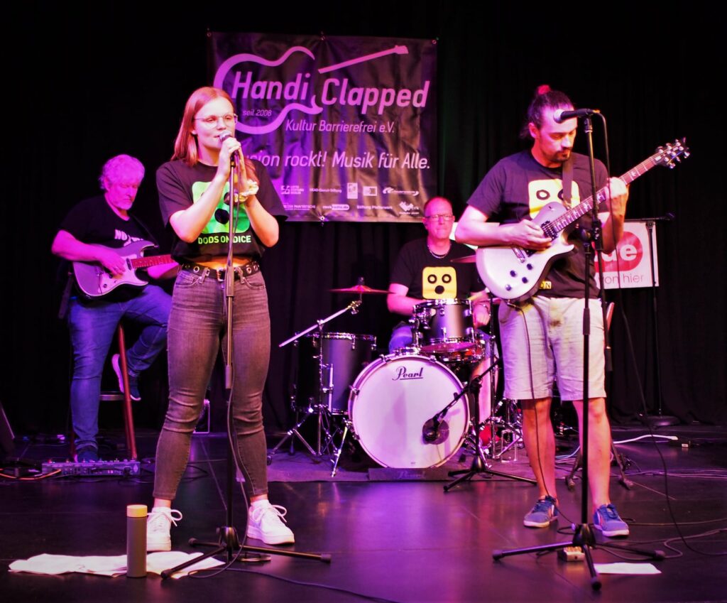 Im Vordergrund eine Sängerin und ein Sänger mit E-Gitarre, beide mit Dons On Dice T-Shirts mit einem Würfel drauf, im Hintergrund ein Schlagzeuger und ein Gitarrist, ein schwarzer Hintergrund mit Handiclapped Plakat