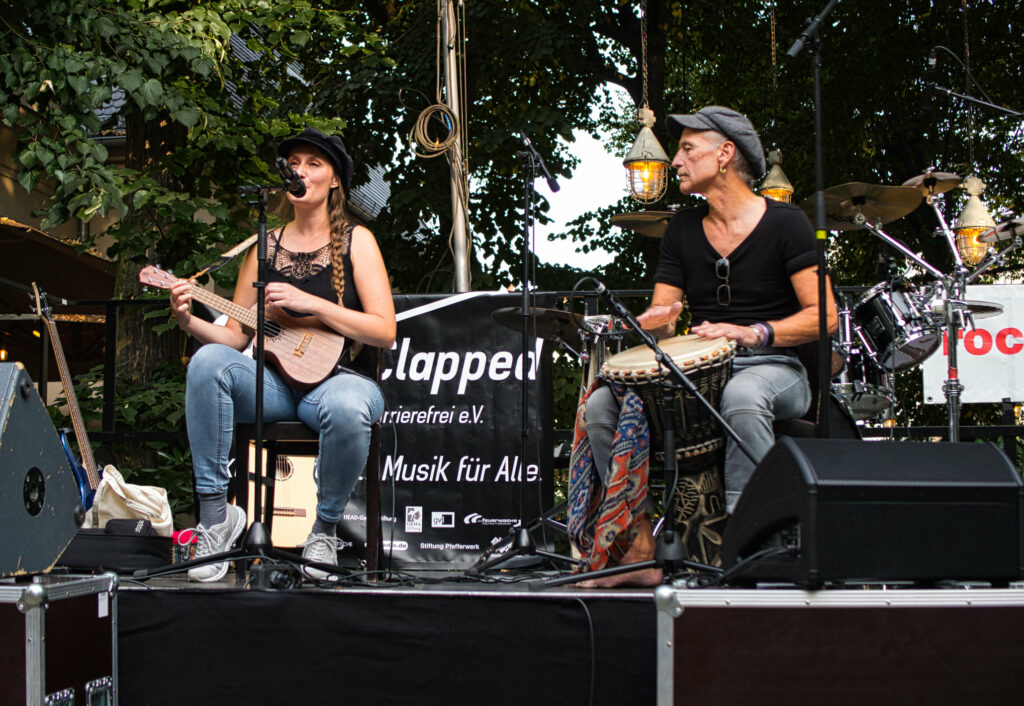 Eine Frau mit einer Mandoline und ein Mann mit einem Bongo spielen live auf der Bühne, im Hintergrund Bäume