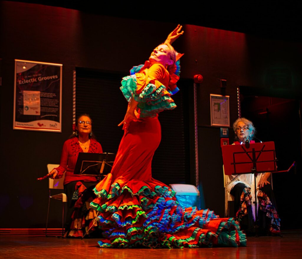 Ein Dame im roten Flamenco-Kleid, mit weiten rauschenden blauen Rüschen an Armen und Beinen, tanzt mit stolz erhobenem Kopf und Arm. Im HInetrgund sitzen zwei weitere Damen, ebenfalls in Flamenco-Kostümen und gucken ihr zu.