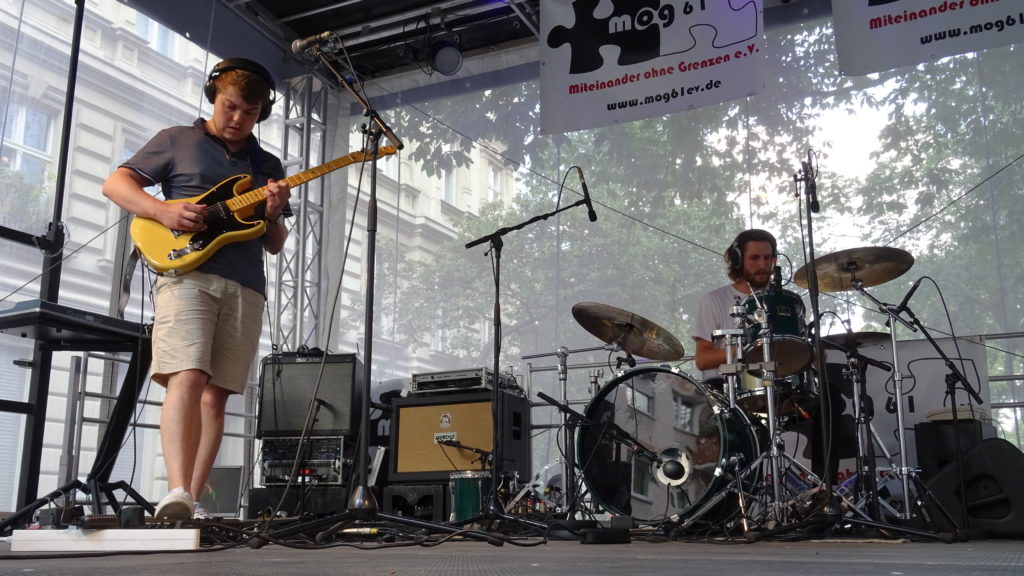 Ein E-Gitarrist und ein Schlagzeuger spielen Live auf einer Bühne, die im Freien steht, im Hintergrund ein mog61 e.V. Plakat