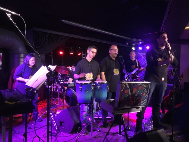 sechs Mitglieder der Band Powerpack live auf der Bühne, mit Bongos, Mikros, Gitarre und Schlagzeug, alle tragen das gleiche schwarze T-Shirt