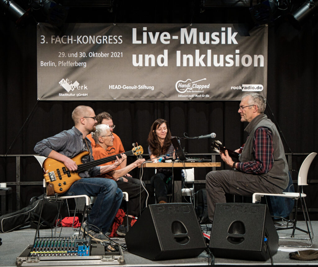 vier Musiker und eine Musikerin sitzen mit zwei Gitarren um einen Tisch auf der Bühne und musizieren zusammen.