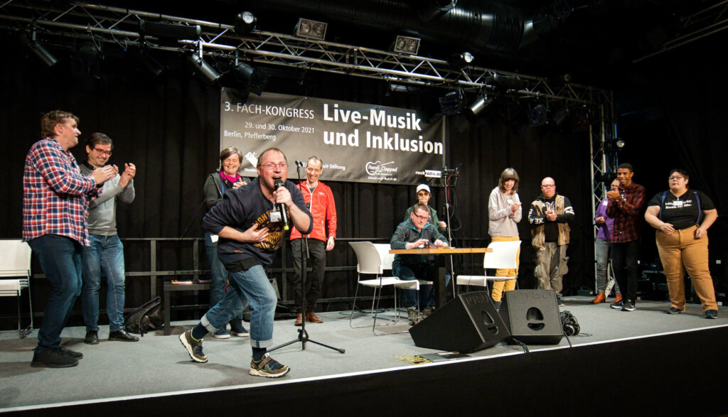 Eine Gruppe von Menschen auf der Bühne, einer singt in ein Mikrofon, einer sitzt am Tisch und bedient ein Tablet