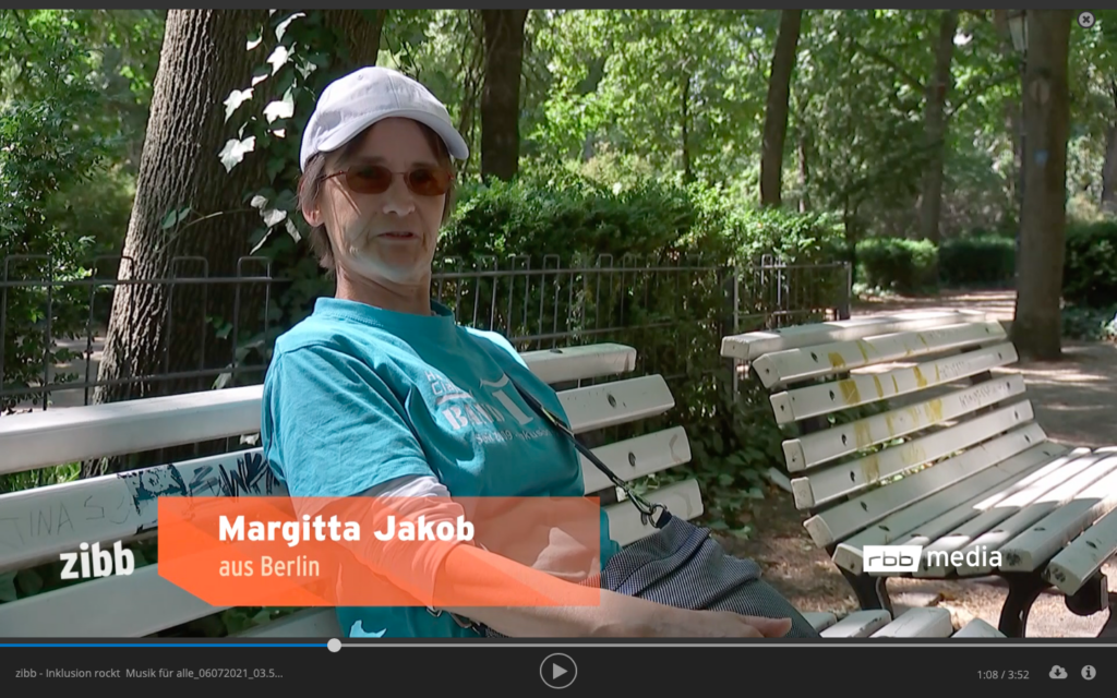 Margitta sitzt auf einer Bank im Park, mit grünem Handiclapped T-Shirt und weißer Kappe. Dazu Schrift Margitta Jakob, zibb Logo und robb Logo