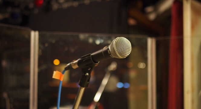 Foto von einem Mikrofon, in einem Mikrofonständer, im Hintergrund unscharf ein Studio.