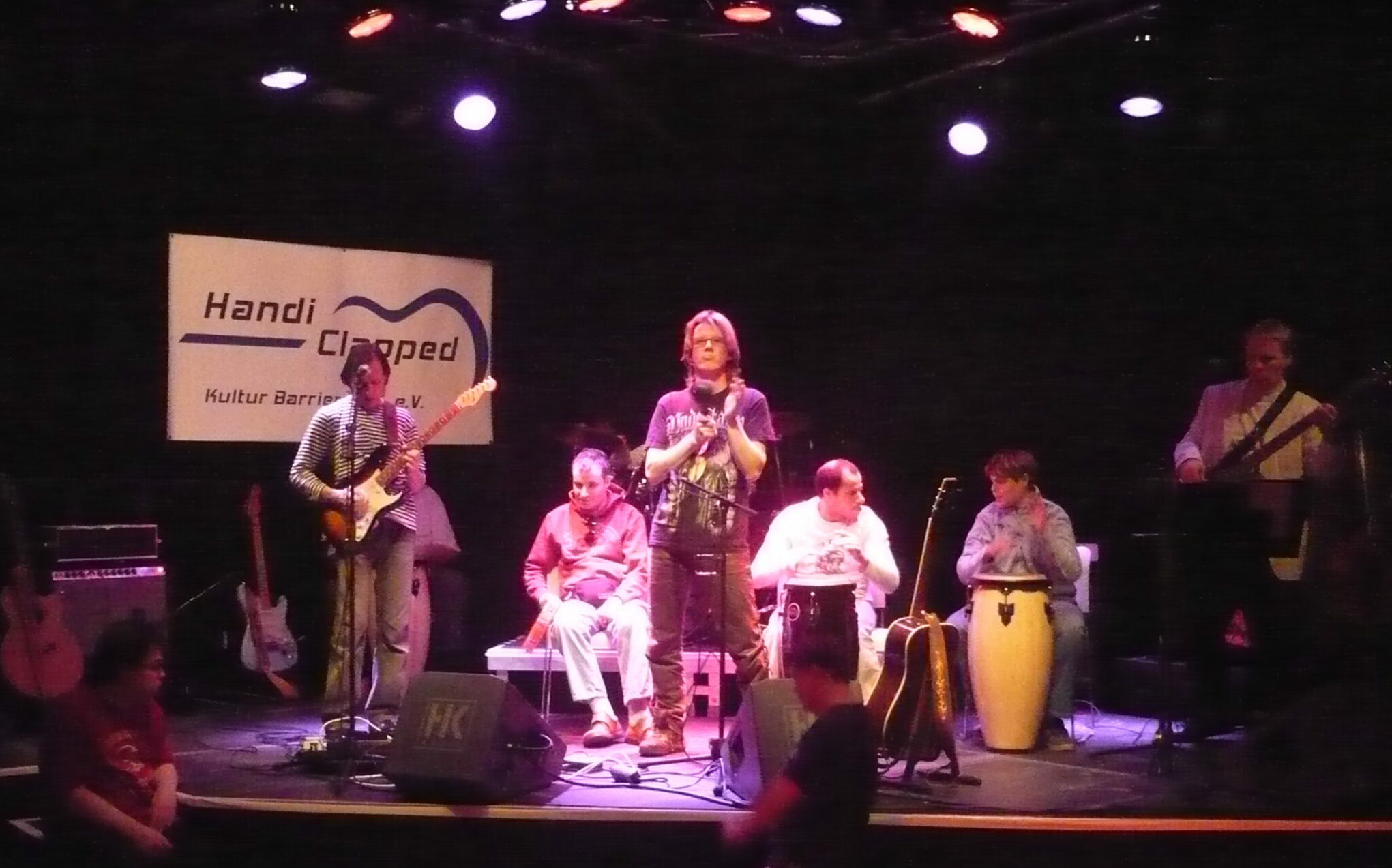 6 Mitglieder einer Band beim Live-Auftritt auf einer Bühne mit Gitarre, Bass, 2 Bongos, einem Schellenbecken und einer Rassel, im Vordergrund 2 Tänzer