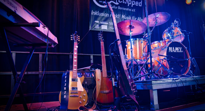 leere Bühne, darauf ein Podest mit Schlagzeug, zwei E-Gitarren und ein Keyboard, im Hintergrund ein Handiclapped Banner