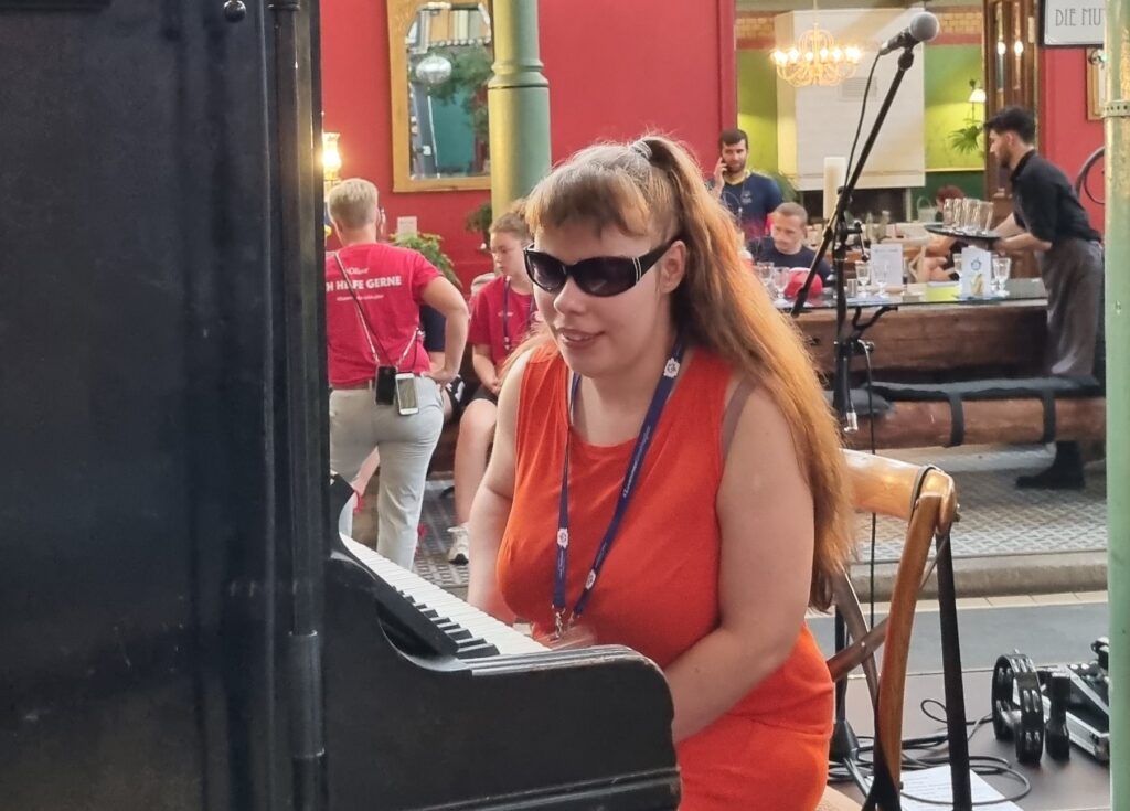 Kevinella mit Sonnenbrille an einem Klavier, im Hintergrund Menschen an einem Bankett