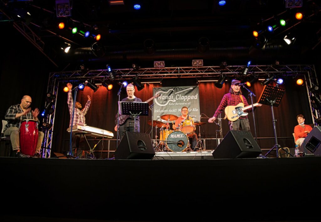 Die 6 Musiker:innen der Band Askanier Rock spielen auf der Bühne ein Konzert. Von links nach rechts: Bongos, Keyboard, Bass-Gitarre, Schlagzeug, E-Gitarre, Bongos. Zum Finale des Songs wirft der Keyboard-Spieler die Hände in die Höhe.