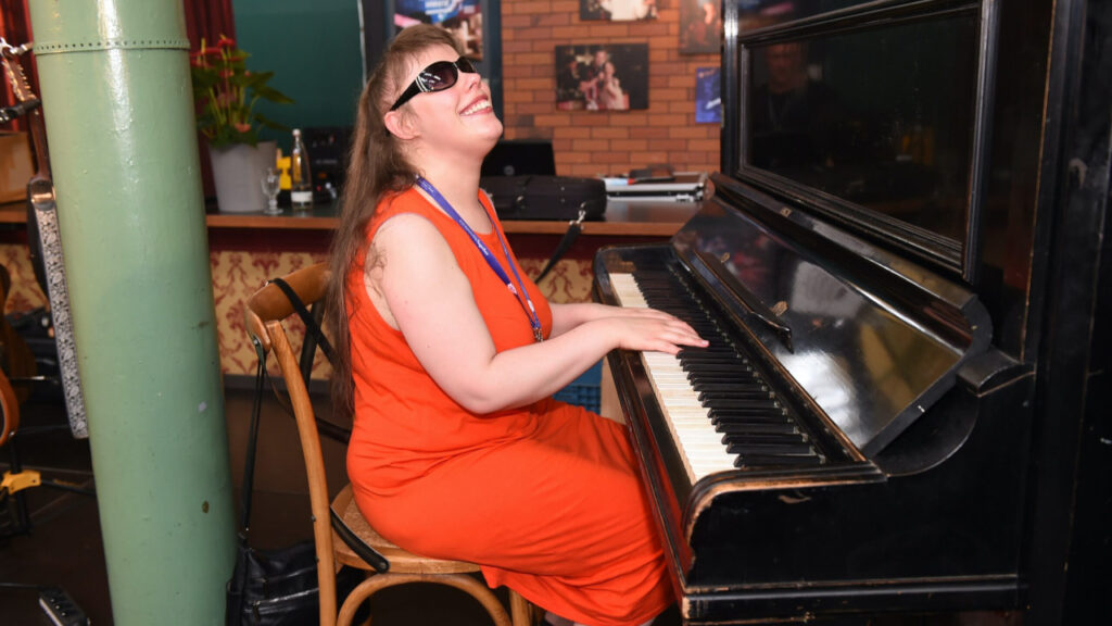 Klevinella, eine bline Musikerin mit schwarzer Sonnenbrille und rotem Kleid sitztam Klavier