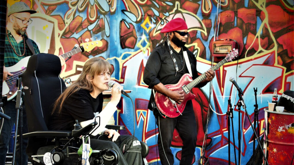 Eine Frau singt ins Mikro, im Rollstuhl sitzend, ein Mann spielt E-Gitarre, beider vor einer bunten Grafitti-Wand