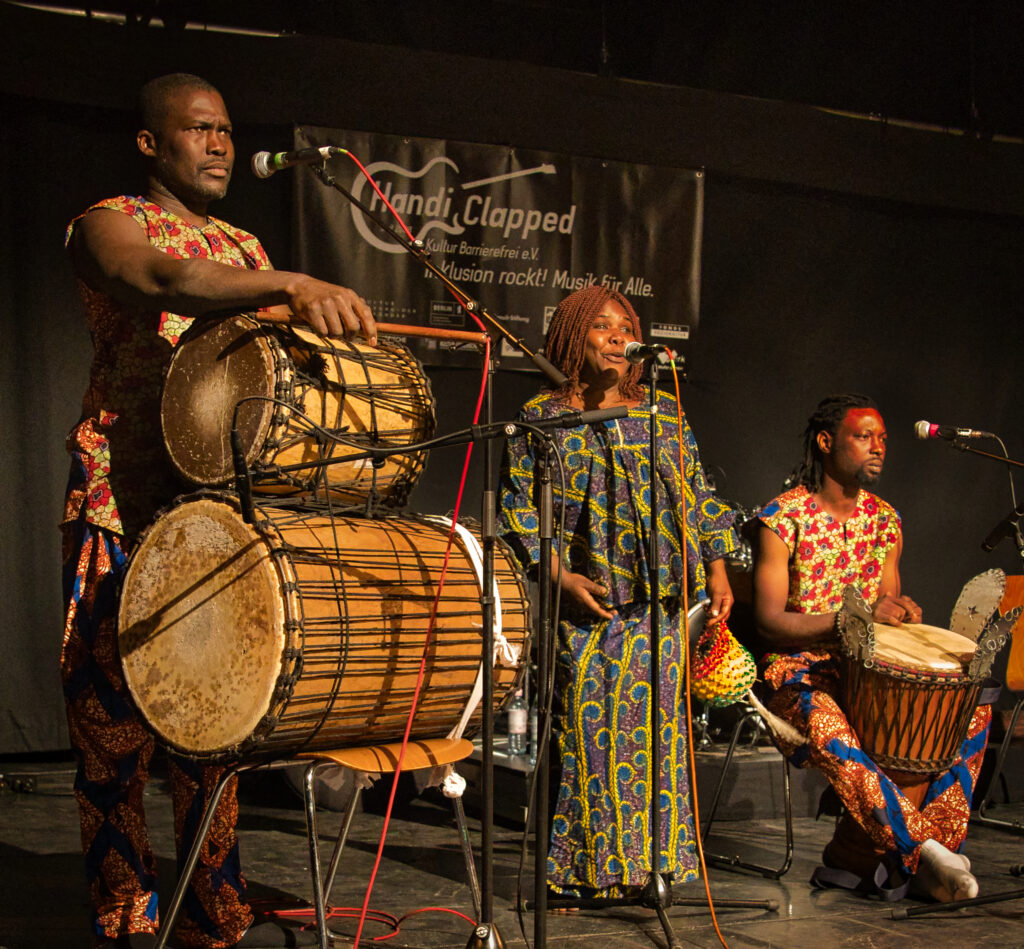 Die afrikanische Band Lanaya auf der Bühne. Man sieht 3 Personen, die Sängerin und Tänzerin in der Mitte, links und rechts je ein Trommler mit verschiedenen afrikanischen Trommeln. Alle tragen bunte, für Westafrika/ Burkina Faso typische, Kleidung.