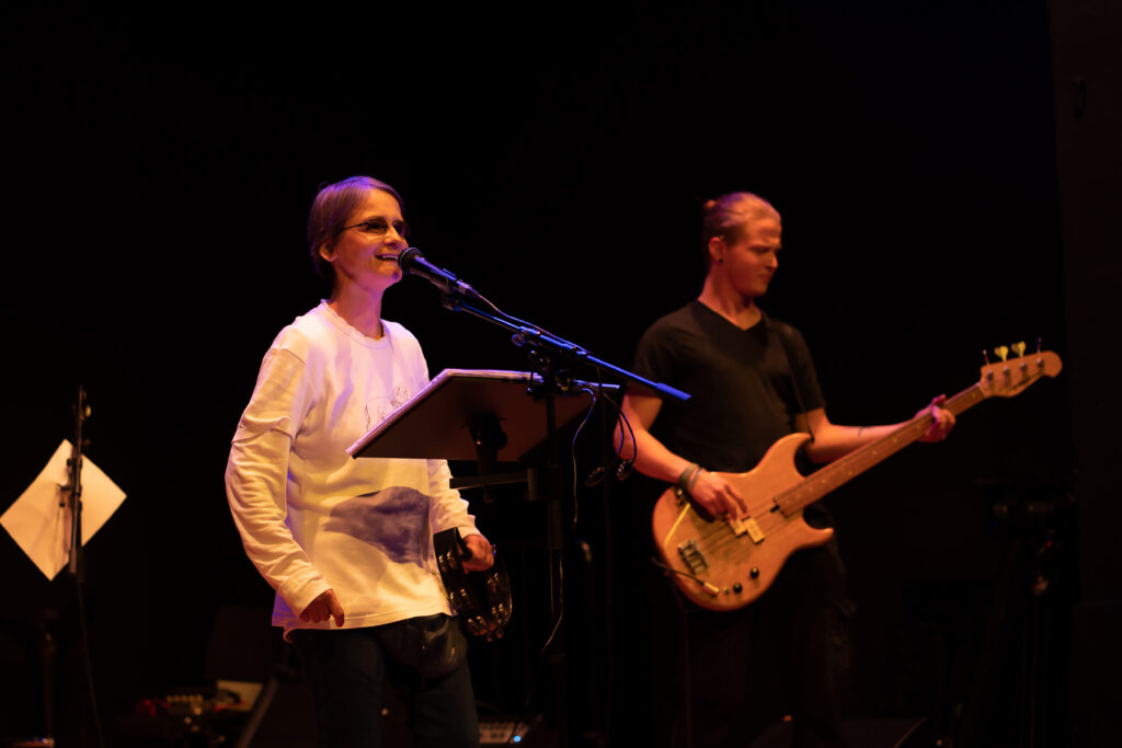 Margitta, die Sängerin von RockAntrieb, steht lächelnd am Mikrofon, rechts von ihr sieht man        im Hintergrund den Bassisten.