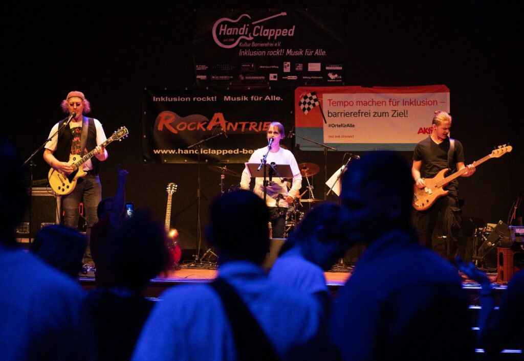 Die Band RockAntrieb steht im Hintergrund auf der Bühne und spielt. Das Foto ist aus dem Publikum heraus aufgenommen , im Vordergrund sieht man mehrere Personen stehen.