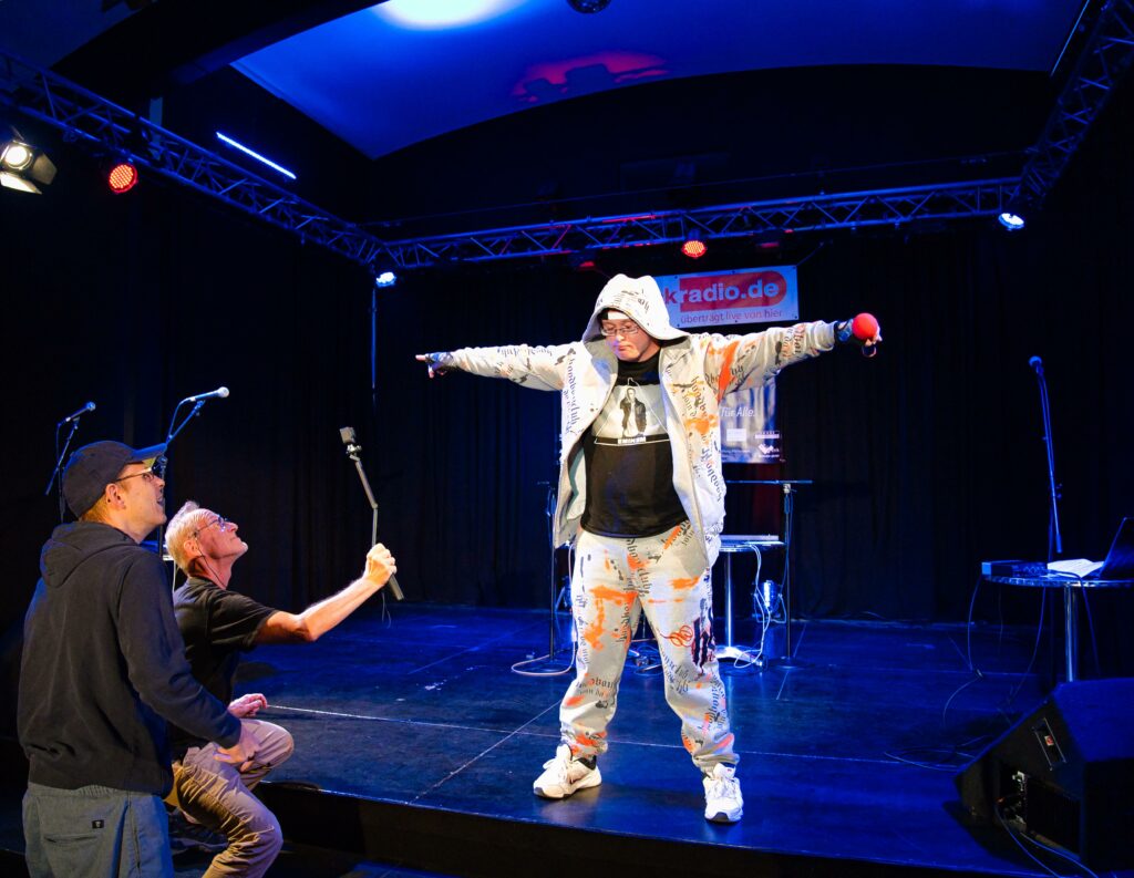 Der Rapper Arthur steht in weißer Kleidung mit Kapuze auf der Bühne und breitet die Arme aus. In einer Hand hält er ein Mikrofon. Vor der Bühne kniet ein Fotograf und macht Aufnahmen, hinter ihm ein junger Mann, der tanzt.