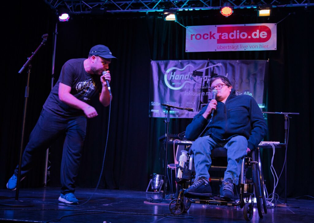 Zwei Rapper auf der Bühne. Einer  beugt sich von links zu seinem Kollegen rüber, der im Rollstuhl sitzt.
