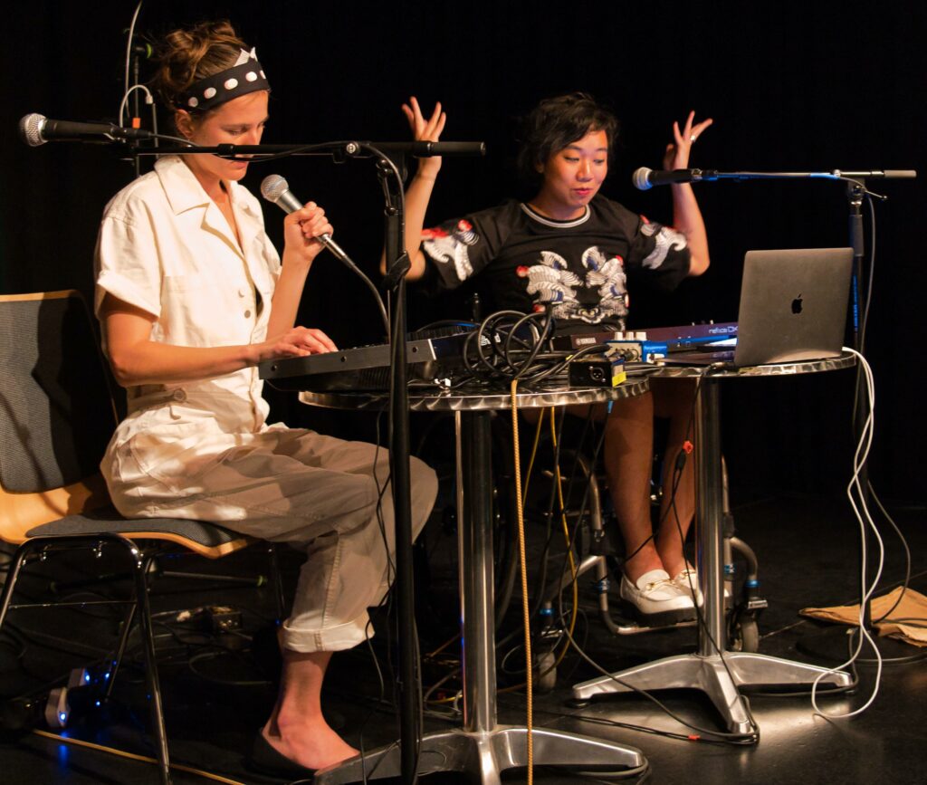 zwei junge Frauen sitzen hinter Keyboard und diversen Computern, sie machen elektronische Musik. Die Frau links im Bild spielt mit einer Hand Keyboard, in der anderen Hand hält sie ein Mikrofon. Die Frau links im Bild sitzt im Rollstuhl und macht mit erhobenen Armen gerade ein tänzerische Bewegung