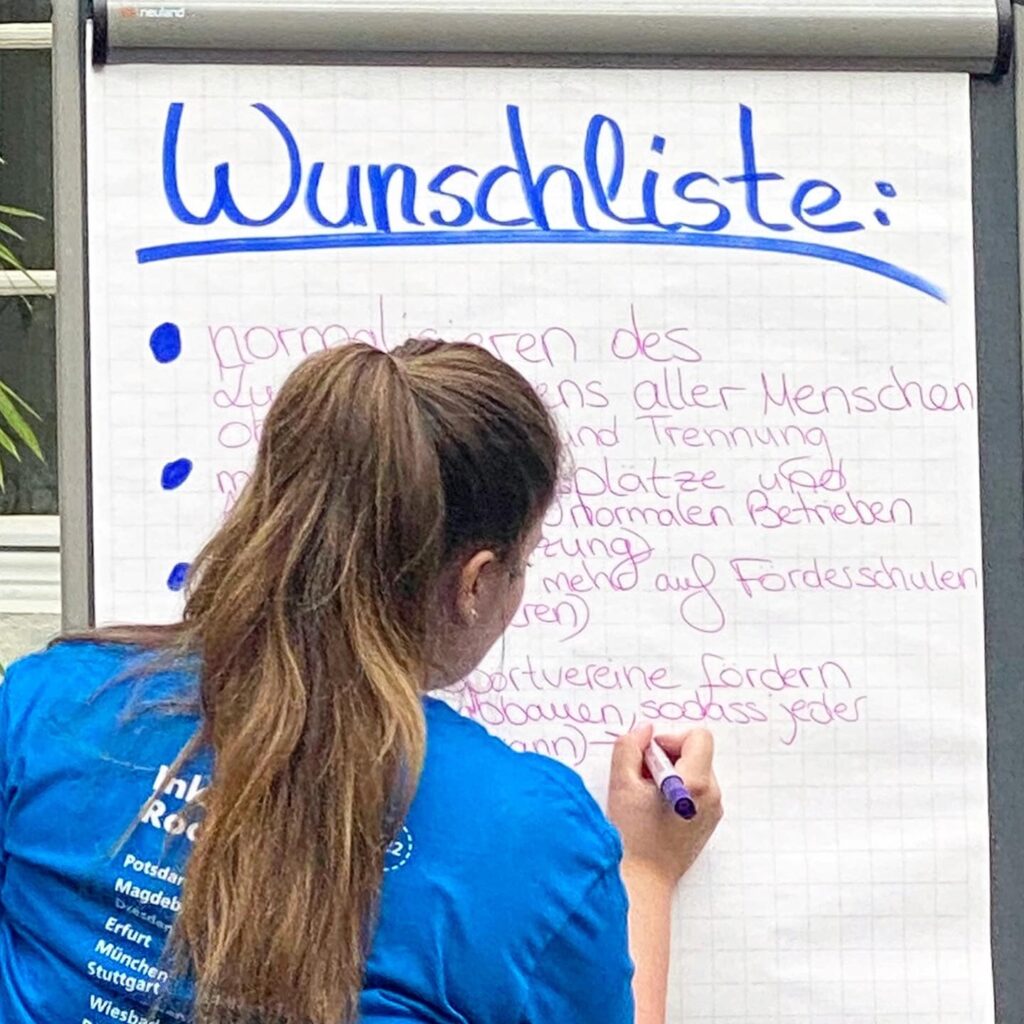 Eine junge Frau notiert Wünsche auf einem Flipchart mit der Überschrift: "Wunschliste". Sie trägt ein azurblaues Event-Shirt mit der Aufschrift: "Inklusion Rockt und Rollt", das von ihrem langen braunen Pferdeschwanz teilweise verdeckt wird.