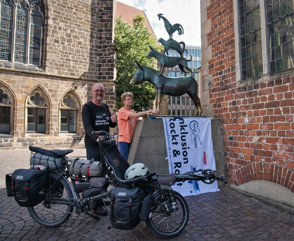 Sven und Sebastian am Denkmal der Bremer Stadtmusikanten. Sebastian erkundet die Skulptur mit seinen Händen. Über dem Sockel der Skulptur hängt die Fahne von  „Inklusion rockt und rollt“.
