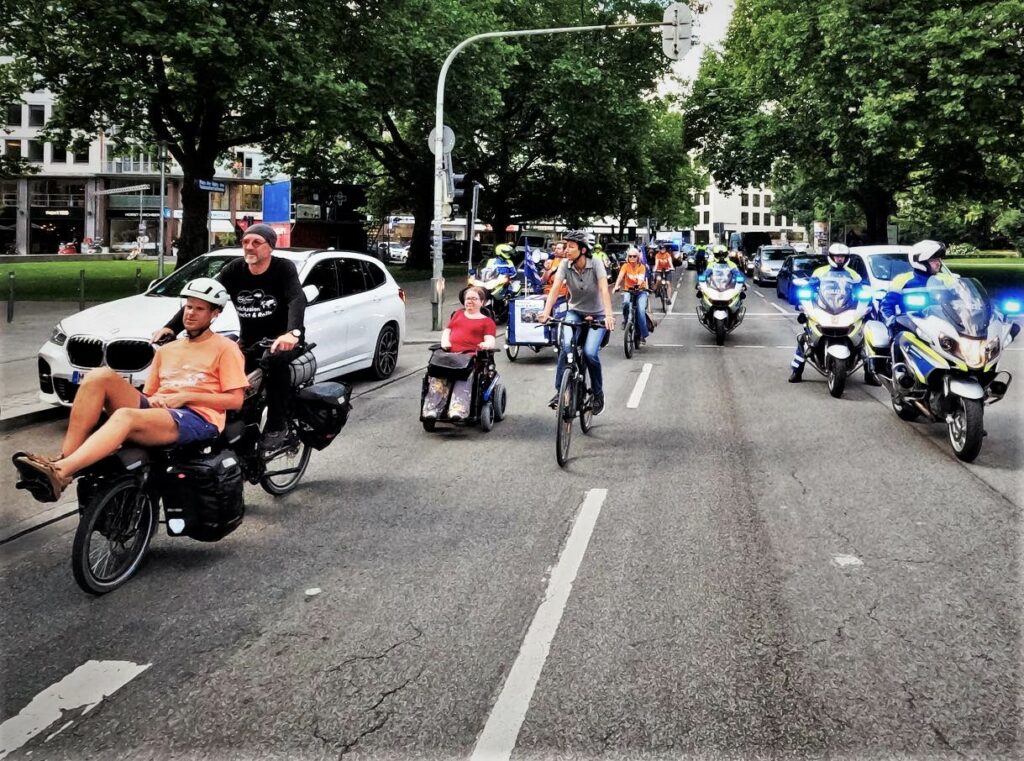 Von der Mitte der Straße aus zeigt das Bild die auf den Kameramann drauf zufahrende Demonstrierende auf Rädern und Rollstühlen. Sie werden von der Polizei auf Motorrädern begleitet.