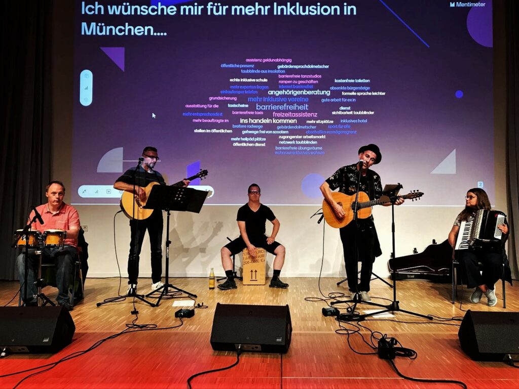 Das fünfköpfige ABM Orchester,bestehend aus Frauen und Männern, spielt auf der Bühne mit Bongos (links), Akustik-Gitarre, Cajón in der Mitte, weiterer Akustikgitarre und Akkordeon auf der rechten Seite. Im Hintergrund ist eine blaue Folie auf die Wand projiziert, mit dem Text: "Ich wünsche mir für mehr Inklusion in München..." Darunter erscheinen Stichpunkte in verschiedenen Farben, z.B. "ins Handeln kommen, Gehwege frei von Scootern, Taubblinde aus Isolation".