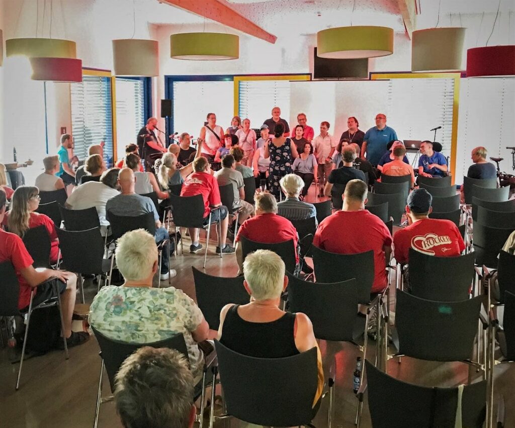 Der 19-köpfige Chor „Wir sind Wir“, bestehend aus einigen Rollstuhlfahrer:innen, singen im Hintergrund vor dem Menschen befüllten Raum.