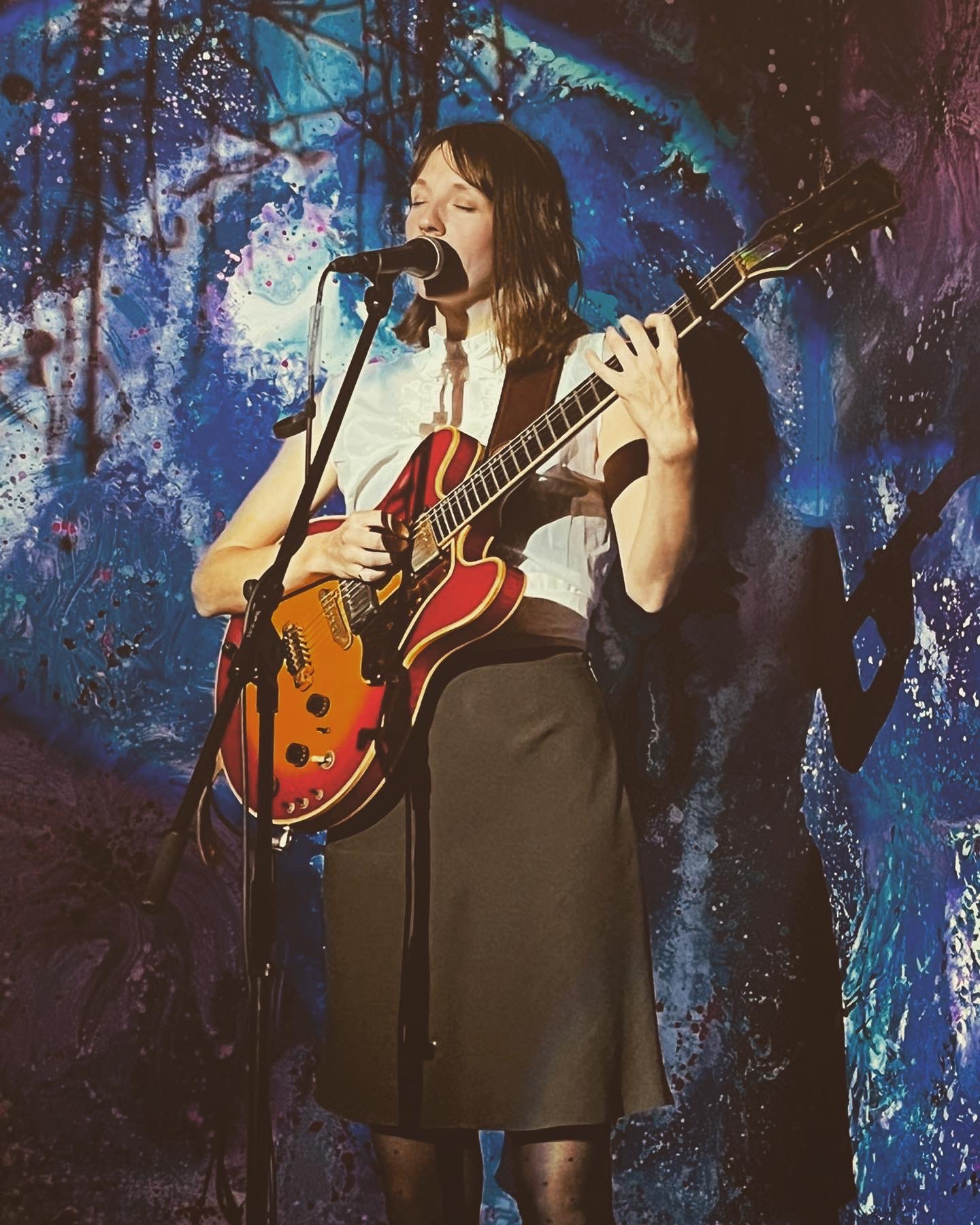 Eine Frau mit Gitarre und Mikrofon, singend vor einer bunten Wand