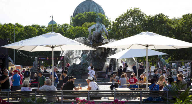 Foto von Menschen an Tischen vor dem Neptunbrunnen, im Hintergrund der Dom, alles bei sommerlichem Wetter