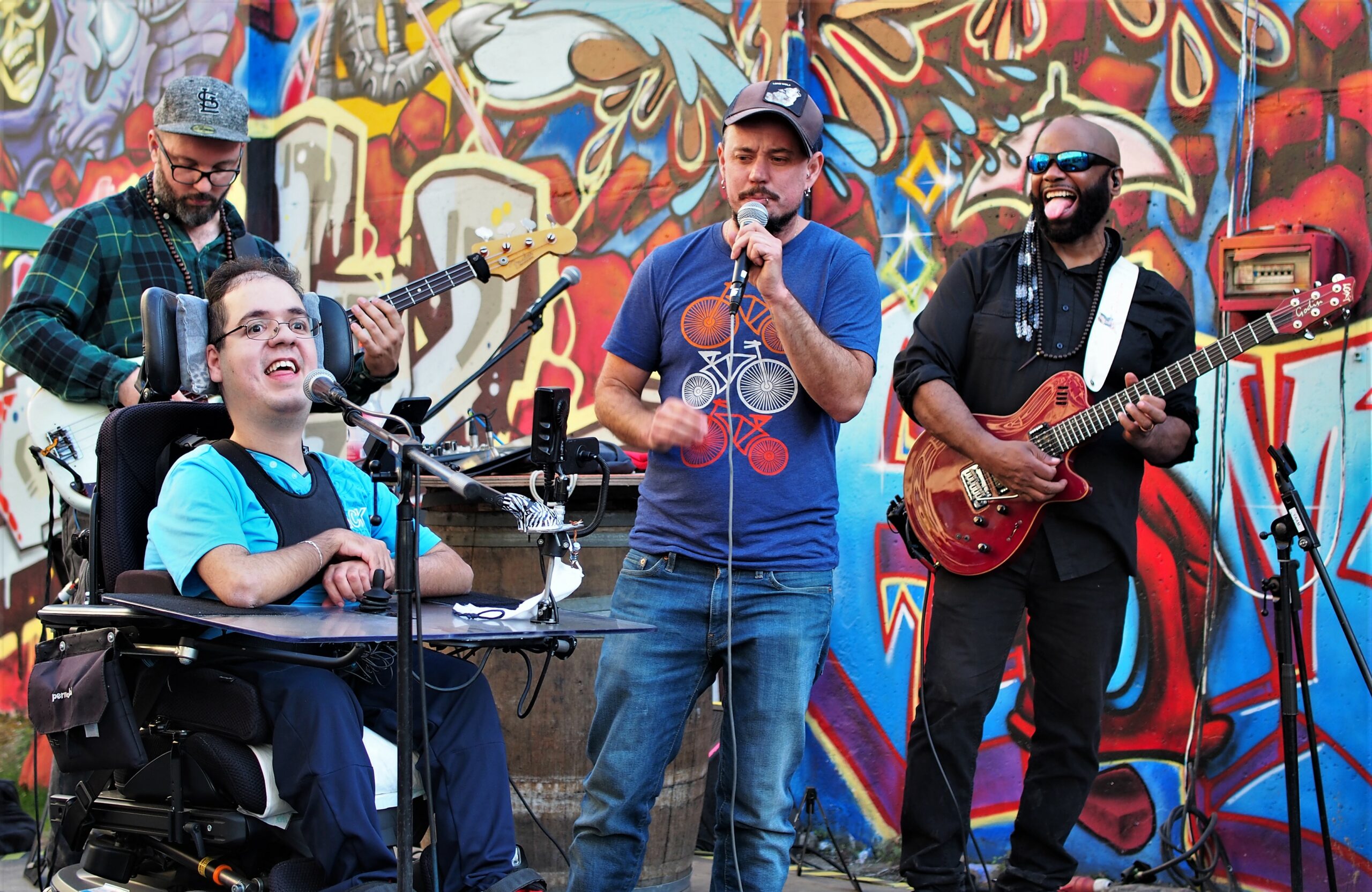 Ein Rapper im Rollstuhl und einer stehend, daneben zwei Musiker und im Hintergrund eine Grafiti-Wand