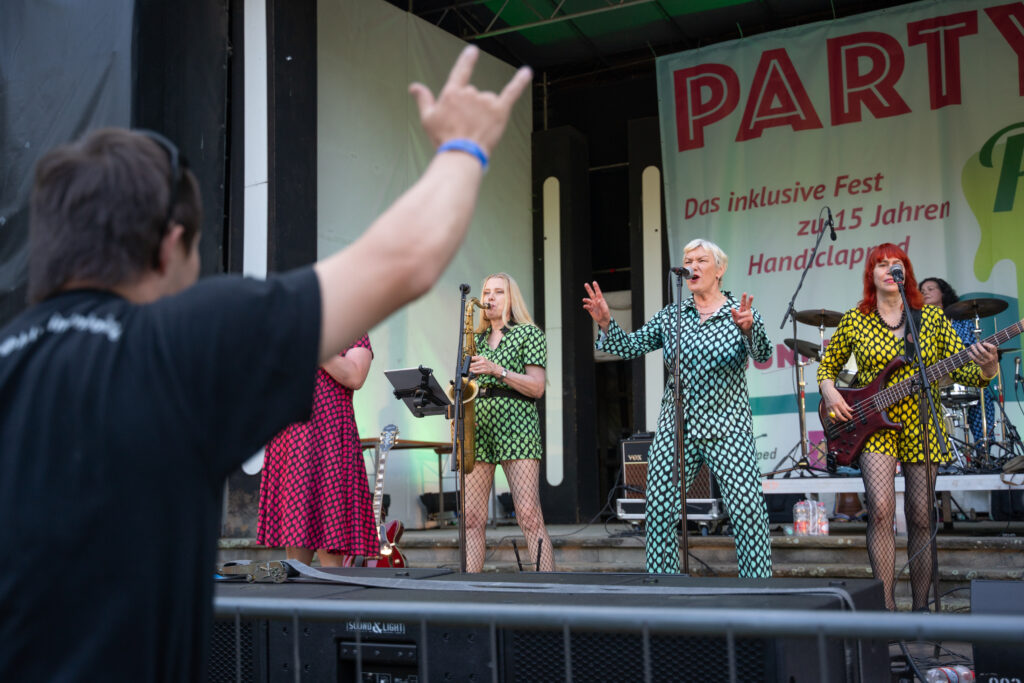 Die Frauenband Die Gabys auf der Bühne beim Fest im Park.  Zu sehen sind eine Saxophonistin, eine Sängerin und die Bassistin. Links im Vordergrund jubelt ihnen ein Fan mit erhobenem Arm zu.