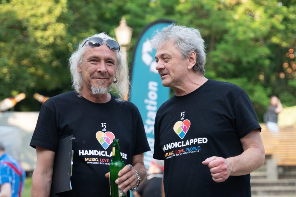 Fest im Park: Handiclapped Mitarbeiter Jens Kaufmann und Projektleiter Peter Mandel unterhalten sich. Sie tragen T-Shirts mit der Aufschrift 15 Jahre Handiclapped, Music.Love.People. Inklusion rockt! Musik für alle