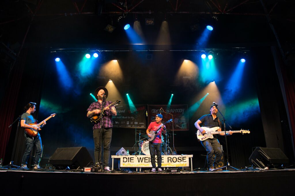Ein Handiclapped Konzert in der Weissen Rose in Berlin Schöneberg, Eine Band auf der Bühne, 2 Gitarristen, ein Bassist, ein Banjo-Spieler sind zu sehen, Bühnenlicht in blau und grün