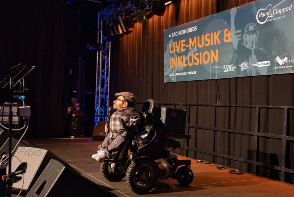 Fachkongress Live-Musik und Inklusion, Raoul Krauthausen im Rollstuhl auf der Bühne, er hält ein Rede. Im Hintergrund das Banner vom Fachkongress.