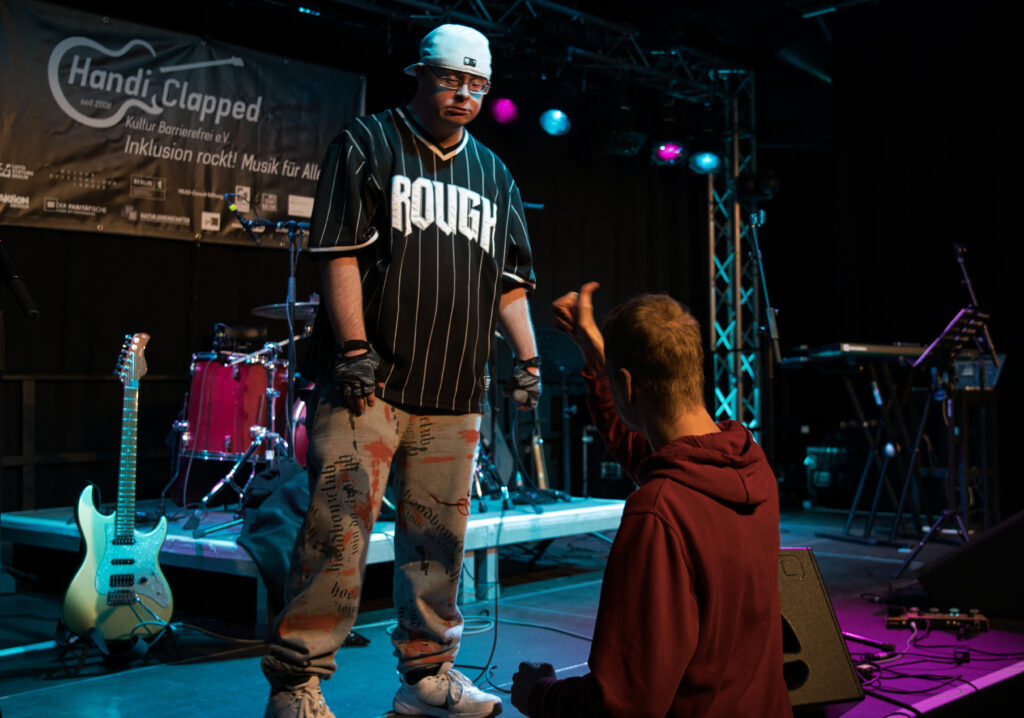 Ein Foto von der Handiclapped HipHop- Offensive. Auf der Bühne der Rapper Arthur mit BaseCap und Handschuhen, direkt vor ihm im Publikum ein Fan der einen Arm oben hat , die Hand mit Daumen oben.