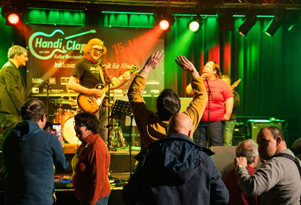 Die Band Die Berfreiten auf der Bühne in grün-orangem Licht, Handiclapped-Banner im Hintergrund. Im Vordergrund, von hinten, tanzen einige Personen, ein von ihnen hebt beide Arme hoch.