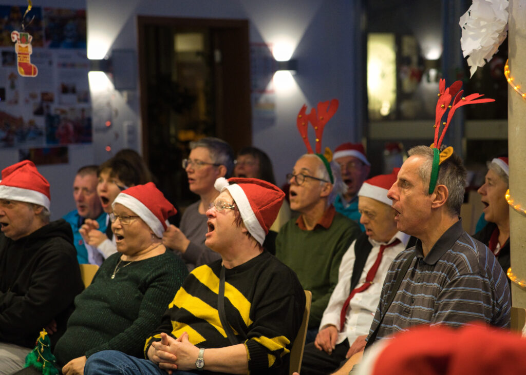 Die Sänger*innen vom Chorworkshop, sie singen mit sichtlicher Begeisterung und tragen alle lustige Weihnachtsmützen.
