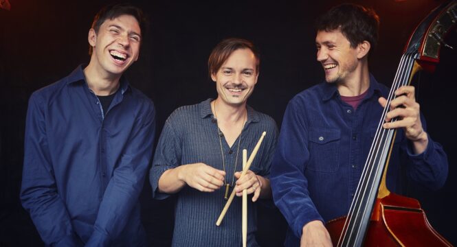 Foto von drei Personen die lachen, eine hält einen Bass, eine andere Schlagzeugstöcke.
