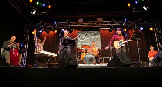 Sechs Menschen als Band live auf einer Bühne innen mit professioneller Beleuchtung und Instrumenten (Schlagzeug, E-Gitarre, Tongas, Keyboard, E-Bass)