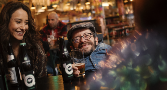 Buntfoto von Raul Krauthausen in einer Kneipe mit fröhlichen Menschen um sich herum und einem Bier von Quartiermeister